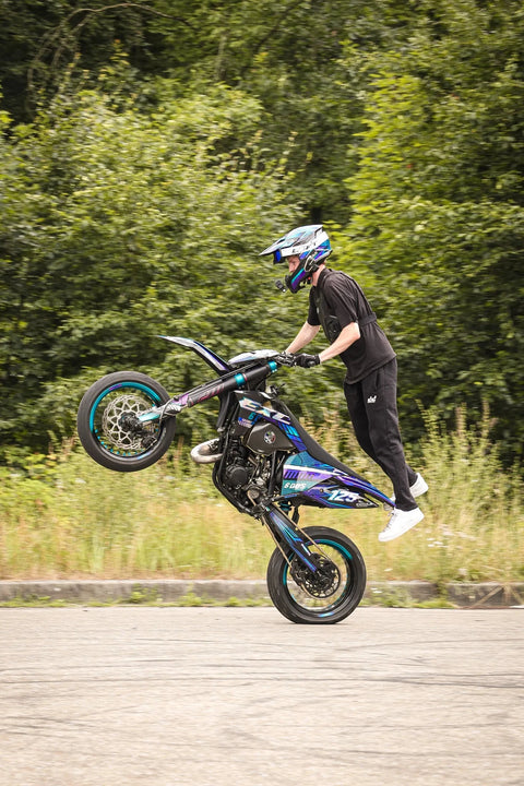 Motard en figure sur une moto à la roue turquoise, devant une forêt