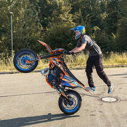 Motard en figure sur une moto orange et bleue, sur une aire bitumée